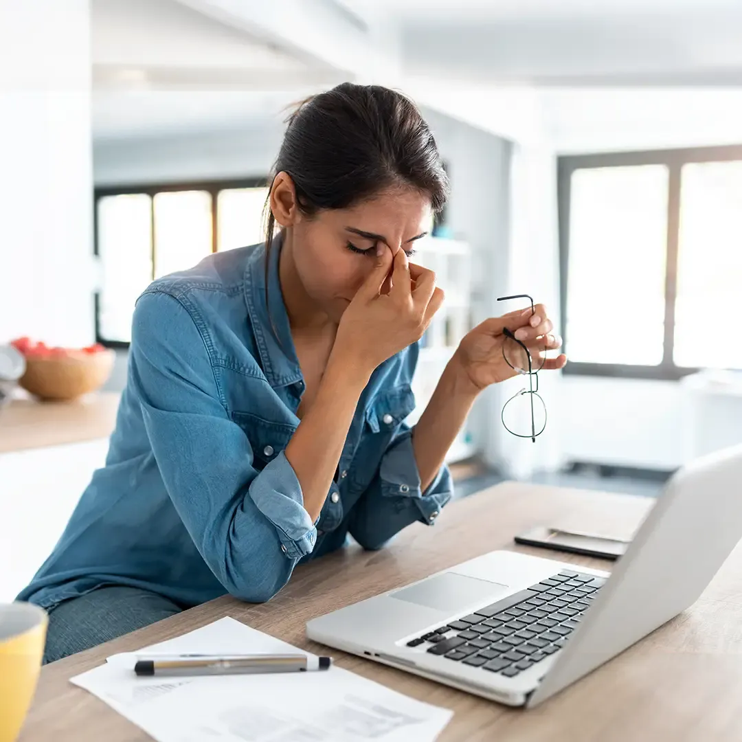 A woman in a denim shirt sits at a table, holding her glasses and rubbing her eyes, looking tired in front of a laptop. Papers and a pen are nearby.