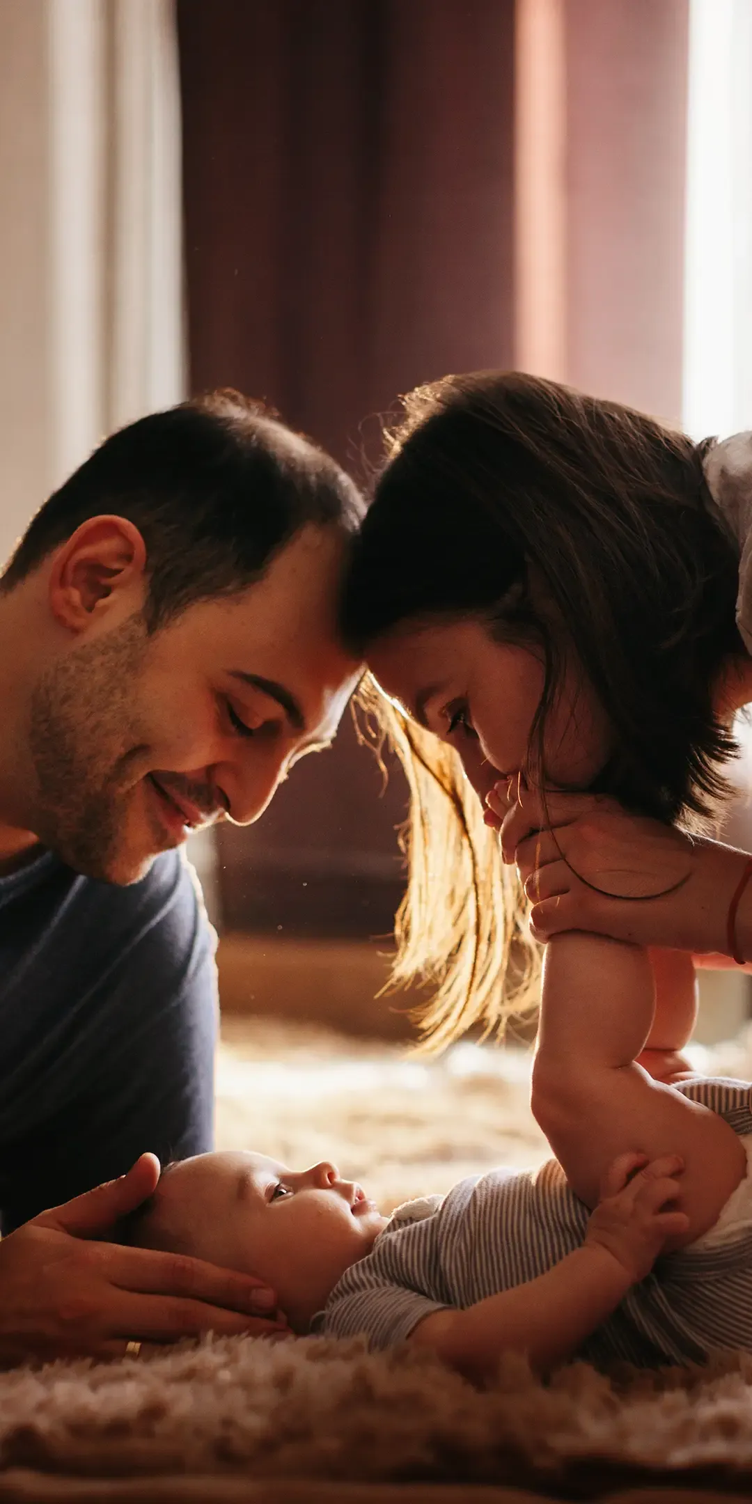 A smiling couple lovingly leans their foreheads together while holding a baby, who is lying down and gazing up at them. Warm lighting fills the room.