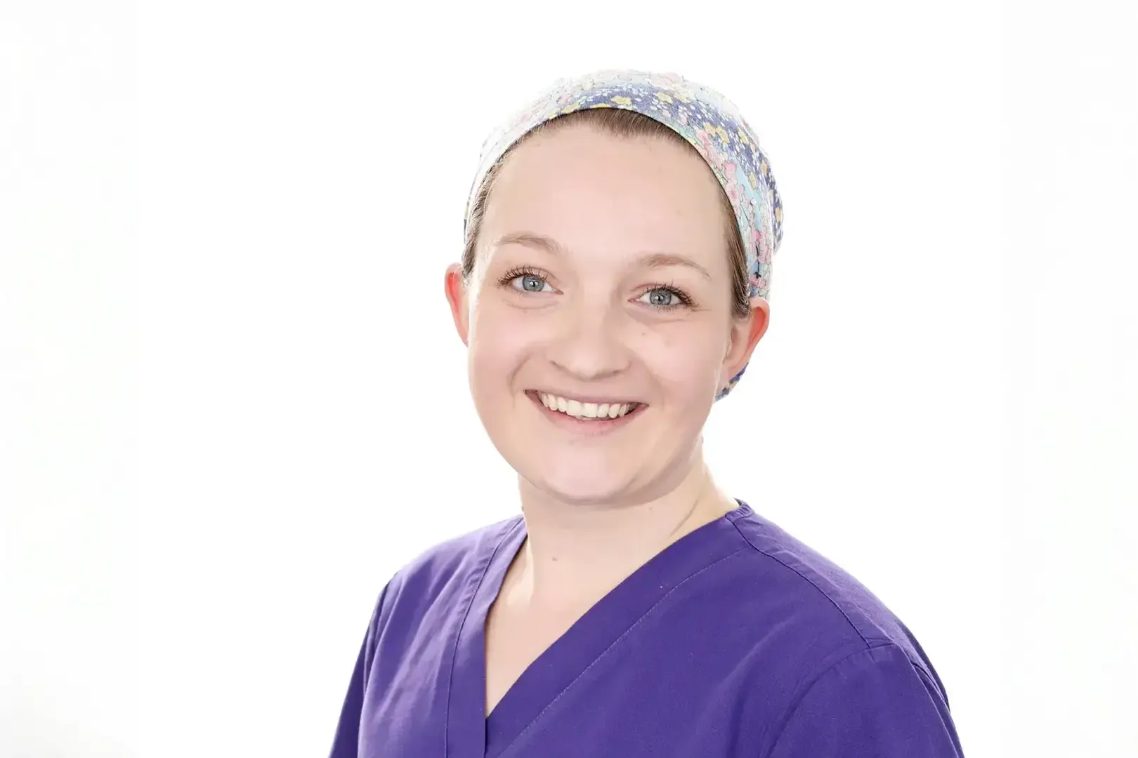 Smiling person wearing a purple scrub top and a floral-patterned headband against a white background.