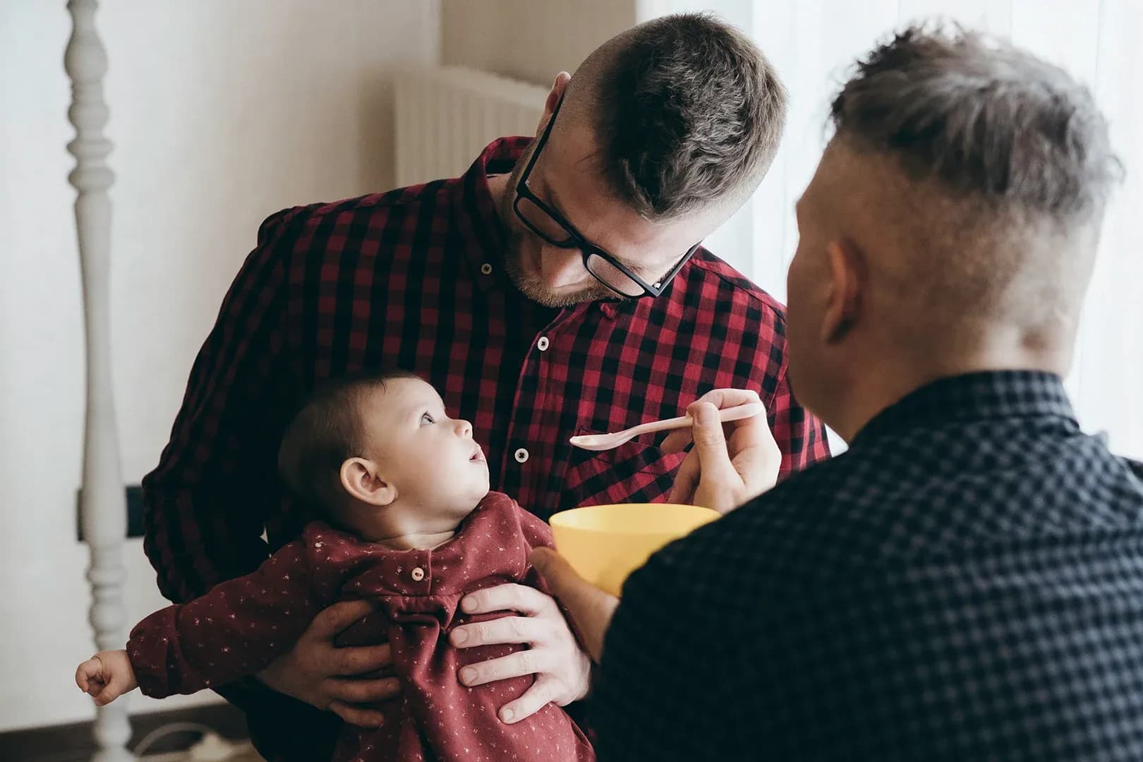 two dads sitting together in their living room, the baby looks up at the dad holding the baby while the other father has a spoon to feed the child
