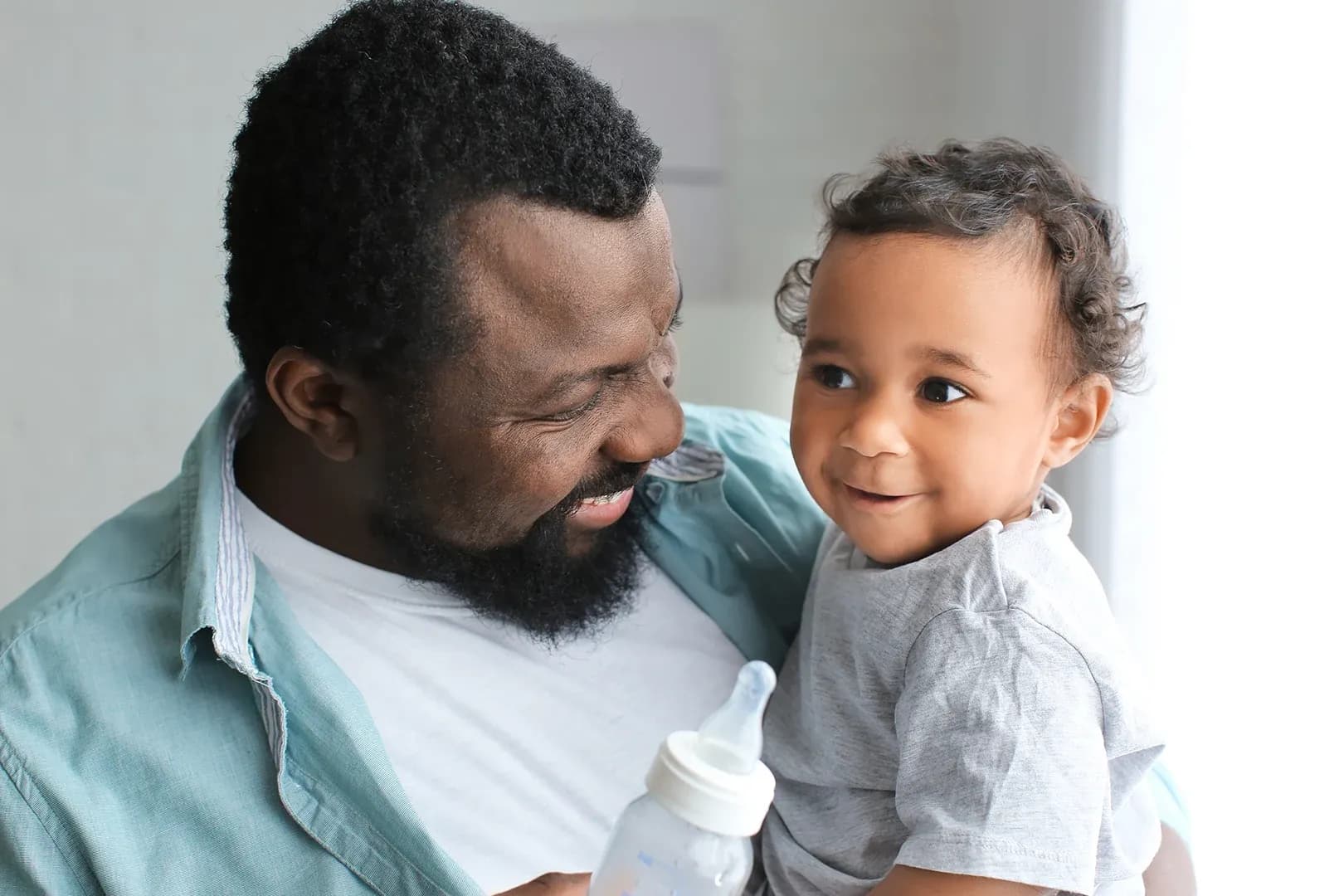 father smiling and looking at young child who is sat in his arms as he tried to bottle feed them