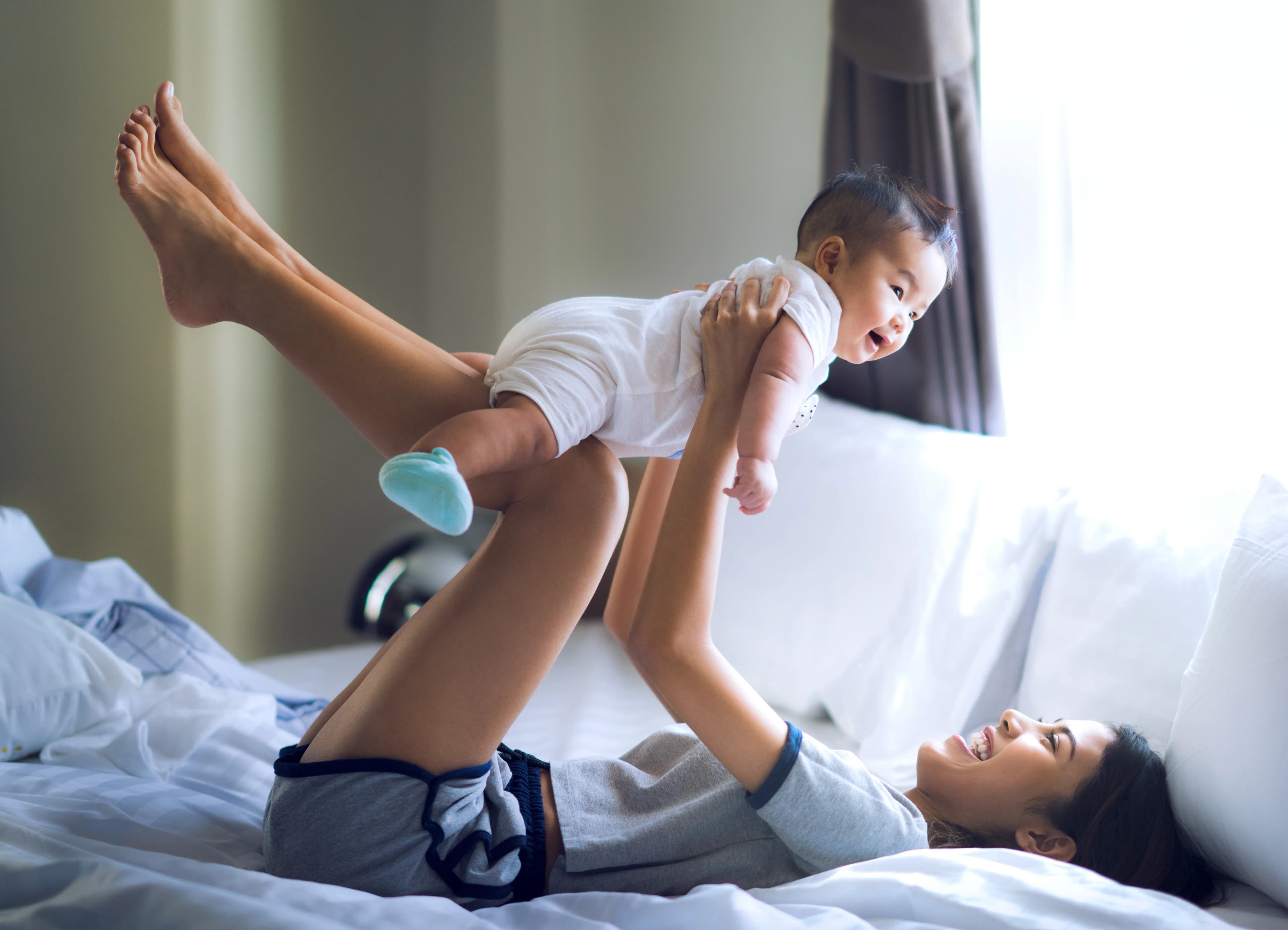 A young women lay on a bed holding her baby above her